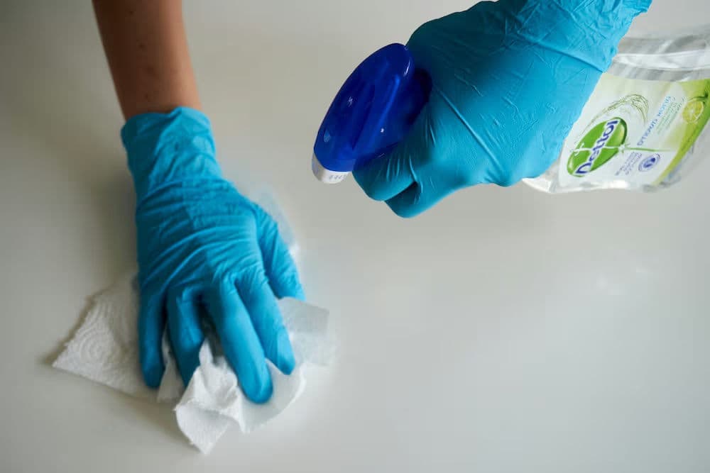 Person in blue gloves spraying a disinfectant while cleaning a surface with a paper towel.